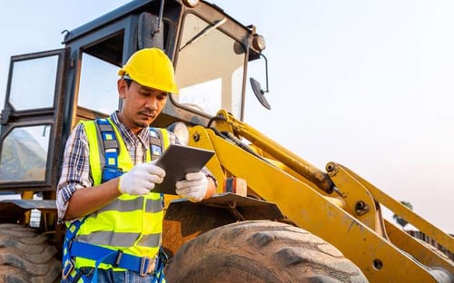 A man with a tablet standing in front of a piece of construction equipment. 